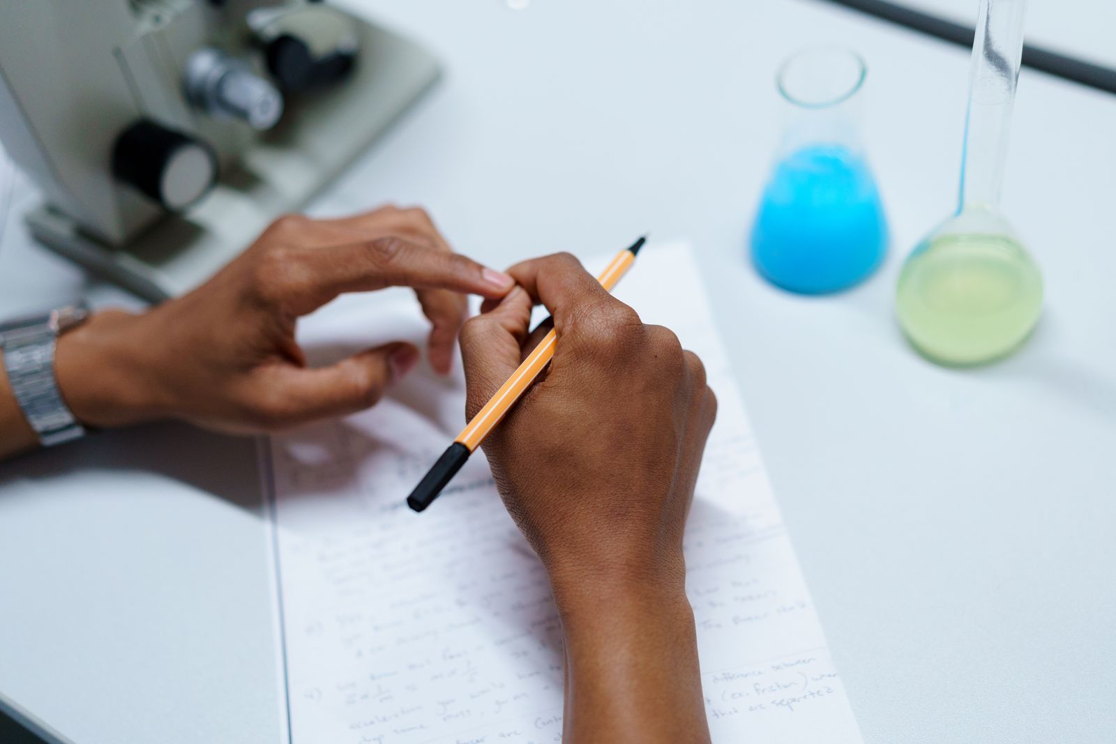 Student writing notes at a desk with two laboratory flasks, studying Cambridge A Level Chemistry 9701